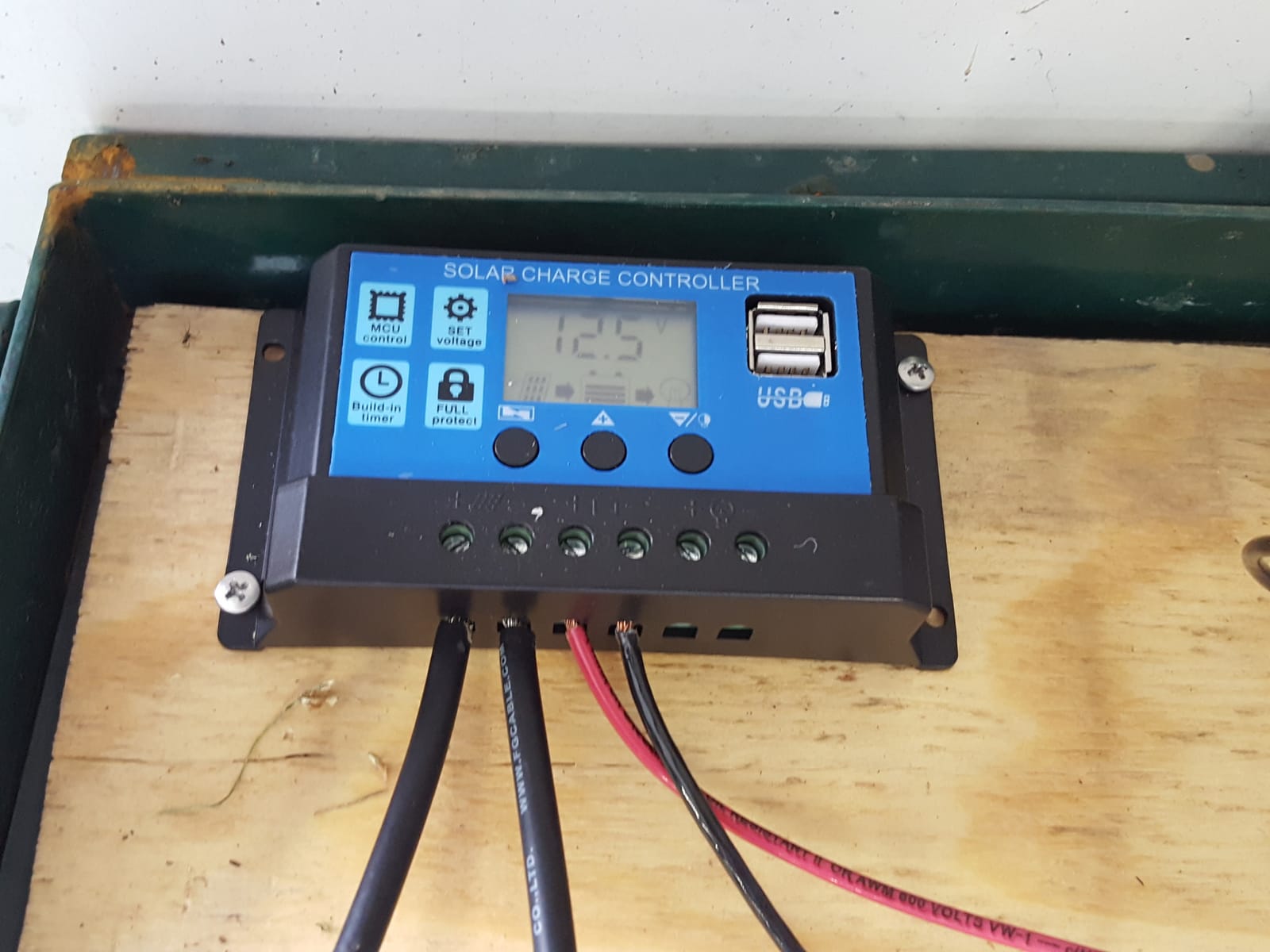 A blue and black solar charge controller screwed to a board, its display showing 12.5 volts, with black and red cables connected to its terminals