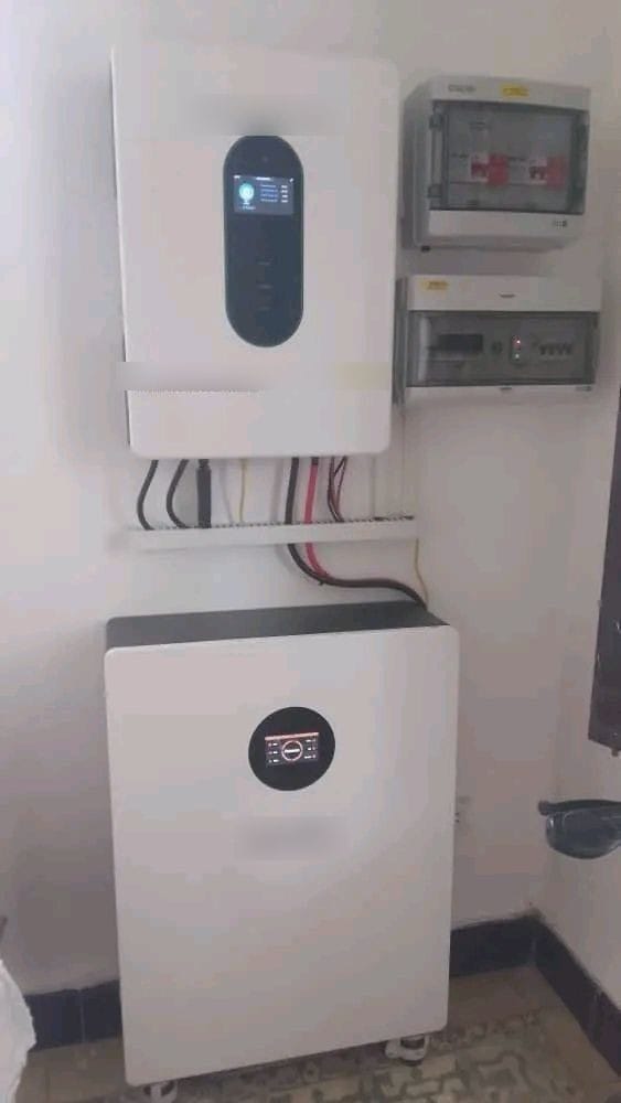 A white inverter on the wall, with two boxes of breakers and protection devices beside it and a lithium battery below, joined by cables