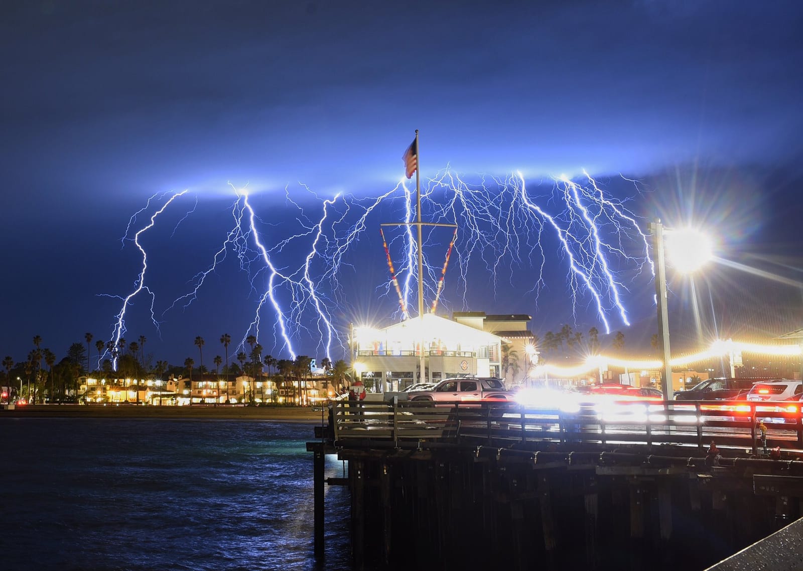 Dozens of lightning bolts crossing the night sky over a lit-up coastal city, seen from a pier