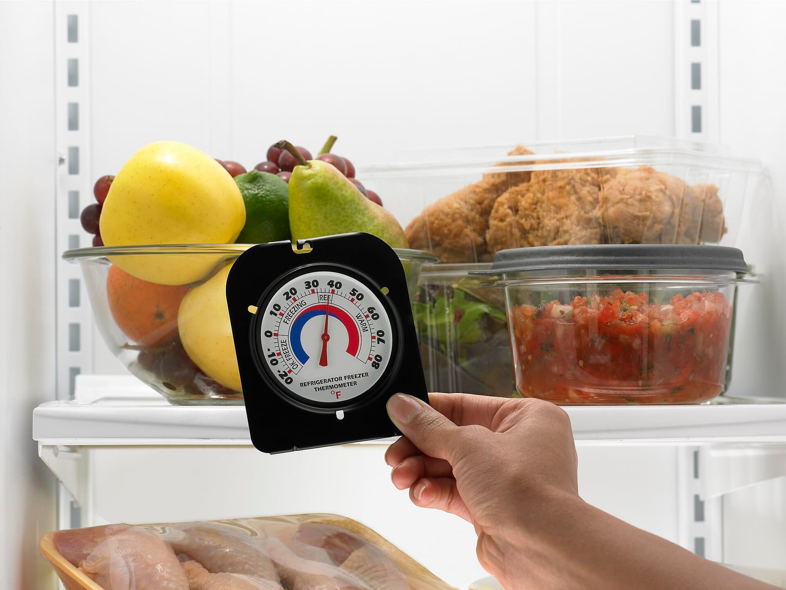 A hand holding a dial fridge thermometer in front of fruit, fried chicken and salsa stored in a refrigerator